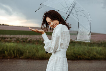 Young woman smiling, holding a clear, transparent umbrella, standing in an open field, checking the...