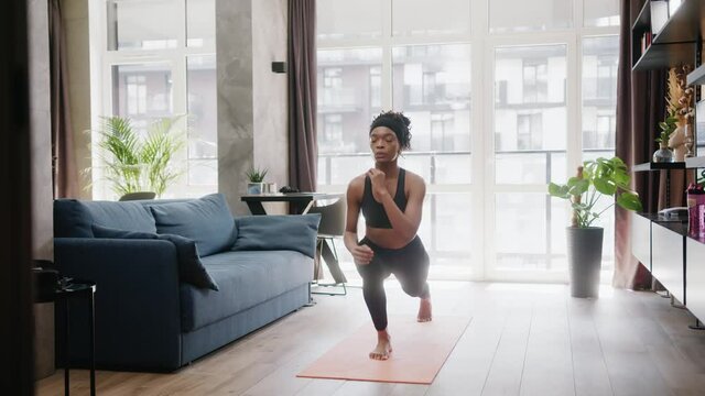 African american fitness woman in sporty bodysuit doing exercises on mat training legs performing lunges squats warming-up in the morning. Sports and healthy life.