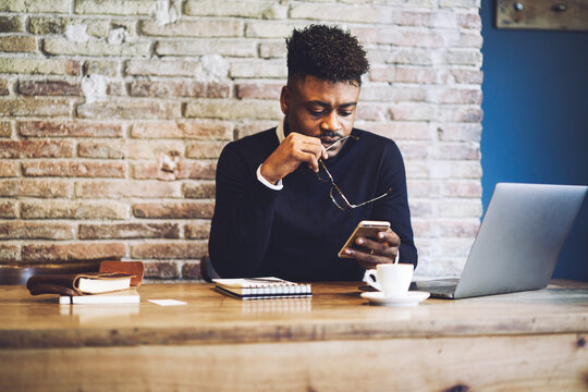 Confident Afro American Worker Of Hotel Reception Waiting For Confirmation Message From Foreign Clients About Confirmation Of Booking While Sitting At Workplace In Modern Interior Using Technology