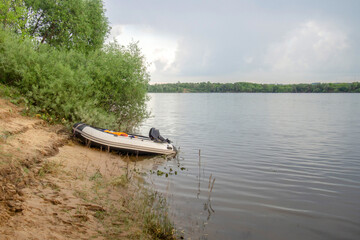 white motorized PVC inflatable boat moored on the sandy shore of the lake against a cloudy sky
