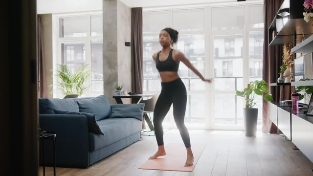 Fit african american sportswoman performing swuats and jumps exercising warming-up cardio training sports routine in the living room. Home isolatio. Fitness. Workout.