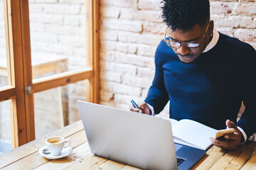 Concentrated afro american businessman working in coffee shop using modern devices and wifi zone while searching more information from different websites and writing important notes in notebook