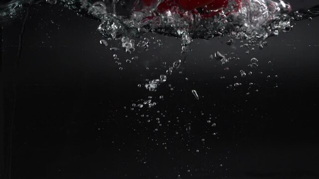 a large fresh red bell pepper falls into the water with splashes, slowly sinking to the bottom, leaving behind a lot of air bubbles. studio shot on a black background. rapid 180 fps