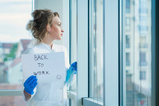 Woman Doctor In White Uniform Showing Paper With Text Back To Work. Pandemic Corona Virus Concept. Female Doctor Looking Out Window