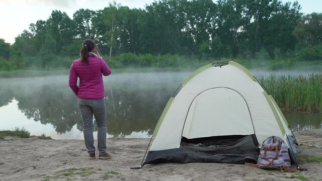 Young Woman Hiker Enjoying View of the Lake Early in the Morning. She is Drinking Hot Tae near a Tent. Vacations and Travel Lifestyle Concept