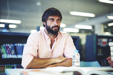 Portrait of young intelligent student sitting at university library table with many text books and...