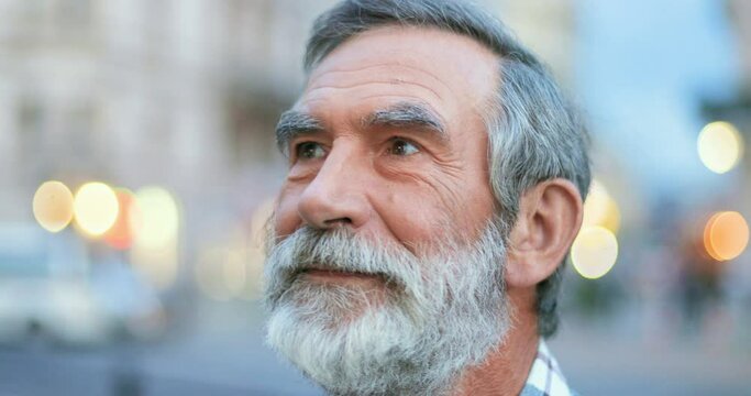 Close up of face of senior Cauasian man with gray hair and beard. Portrait of handsome old grandfather looking at side ad then turning face to camera with smile at street in town. Outdoors.