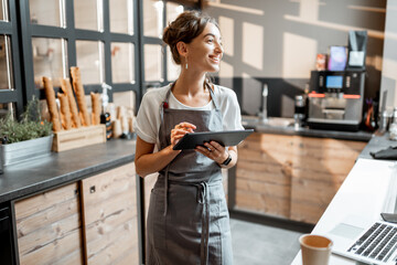 Young saleswoman working with a digital tablet at the counter of cafe or confectionary shop....