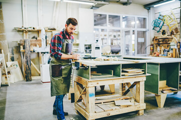 Young handsome male owner of manufacturing and crafts business working in his workshop with professional tools and equipment, serious foreman repair wooden furniture using artisan instruments