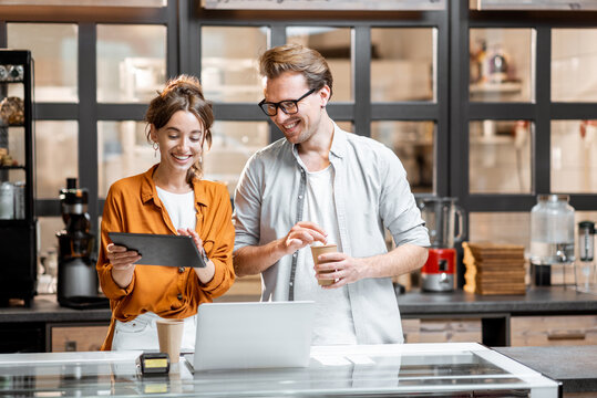 Two young managers or shop owners having some discussion while standing with a digital tablet at the counter of the shop or cafe. Small business management concept