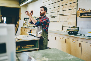 Serious bearded man spending time on hobby making diy furniture using timber and equipment in garage, skilled caucasian male owner of manufacturing workshop looking at wooden detail for construction.