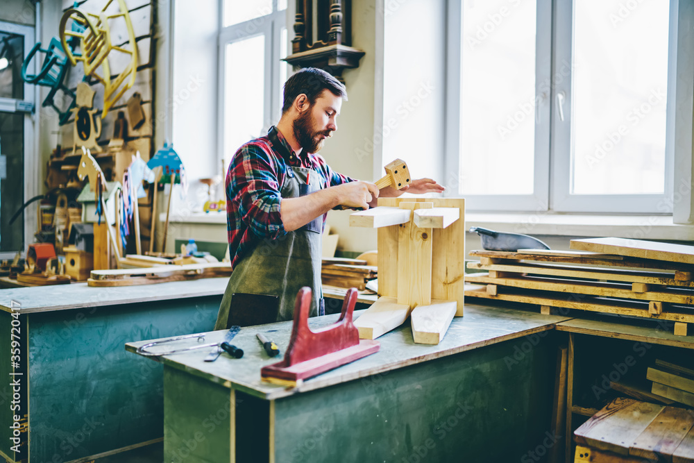Poster serious skilled man in apron working on handmade stool using instruments in his garage spending time