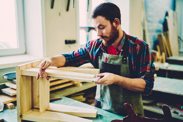 Handsome bearded man working with wooden construction in own small-sized furniture company, Caucasian talented male craftsman engineering decorative equipment with tools during handmade hobby