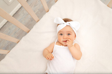 portrait of a baby girl 6 months old with a bow in the crib in the children's room in white clothes and looking at the camera, baby's morning, baby products concept