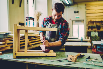 Caucasian craftsman improving decorative chair during woodworking hobby in manufacturing restauration, 30 years old male worker in apron concentrated on handmade equipment during engineering