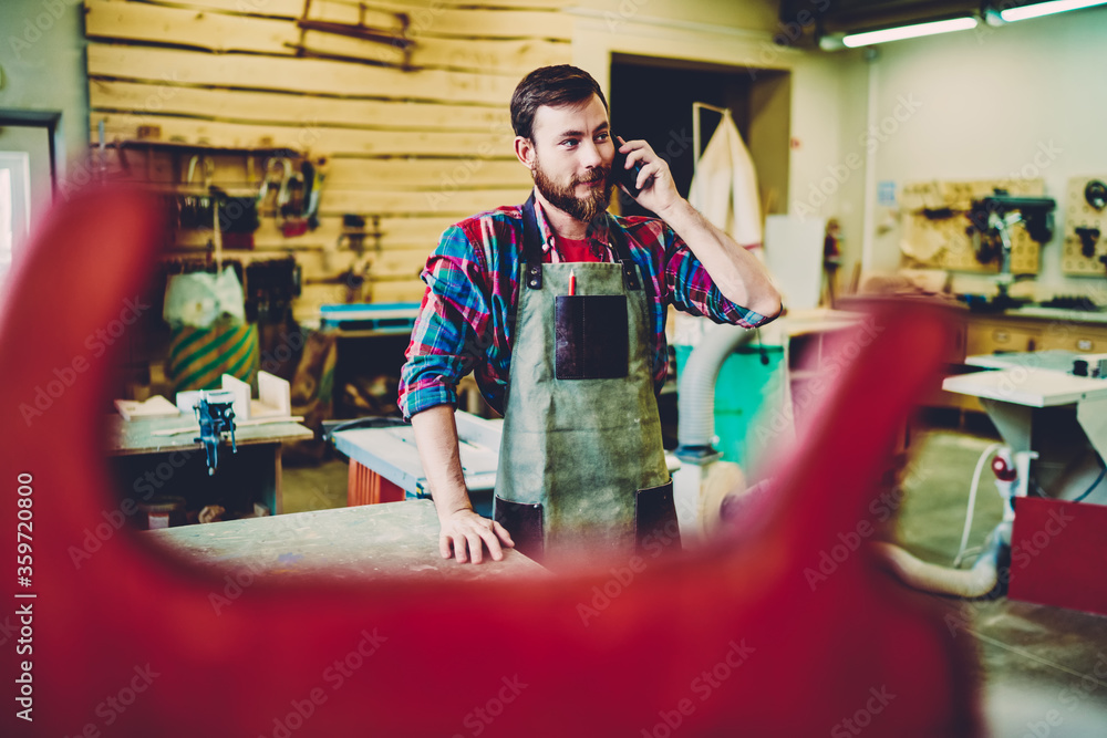 Poster positive bearded male joiner talking on mobile phone during working day in own workshop with equipme
