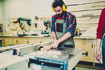 Professional male carpenter in eyewear and headphone for protection saw wooden plank for furniture construction, caucasian craftsman using equipment in workshop making wooden details himself.