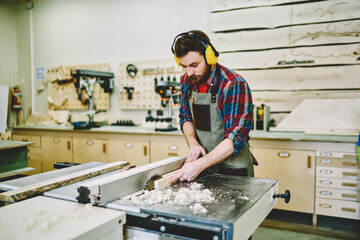 Concentrated young man in electronic headphones for noise cancellation in own workshop using circular measurement for improving wooden plank, serious male carpenter making carving with timber