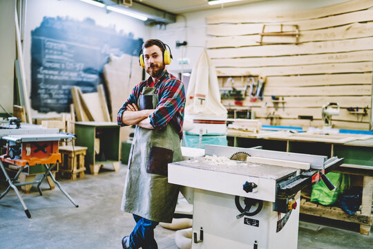 Half length portrait of self-employed woodman in work apron standing near lathe instument with crossed hands and looking at camera, successful male woodworker in protection headphones posing indoors