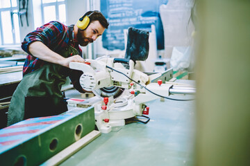 Caucasian professional craftsman concentrated on carving construction indoors using electronic circular machine for decorative wooden plank, bearded woodman in protective headphones improving details