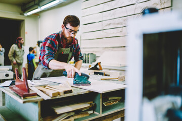 Serious bearded male in eyewear for protection working with electric instrument for polish wooden plank , skilled foreman making details for furniture prepare surface with grinder machinery.