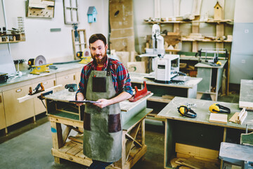 Portrait of cheerful bearded male owner of craft workshop standing near woking place with notepad for organizing job, smiling businessman in apron looking at camera checking clients ordering  .