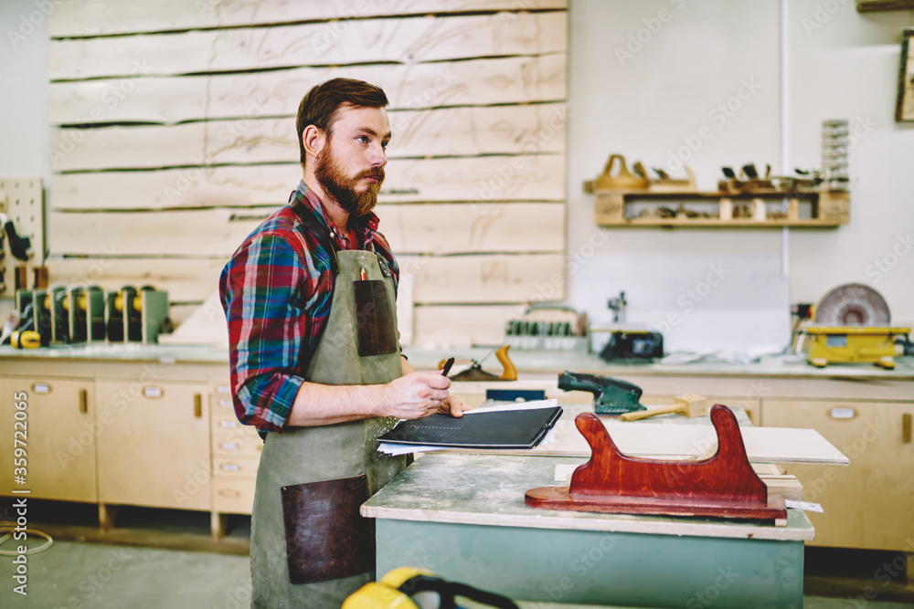 Poster pensive bearded handyman in apron standing with notepad pondering on organizing day in workshop, ser