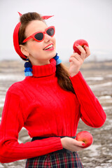 Portrait of a young beautiful girl in a bright red sweater, sunglasses and blue earrings with two red apples in their hands. Beauty, fashion.