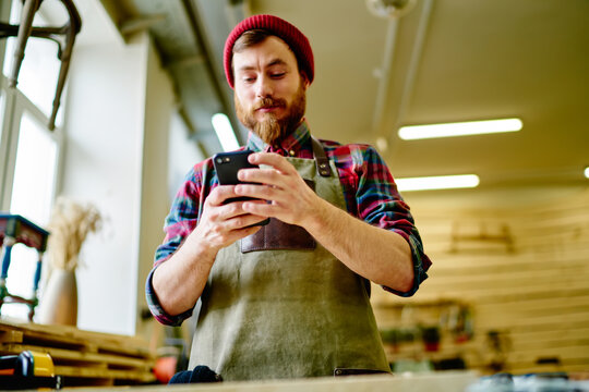 Calm thoughtful craftsman using smartphone in workshop