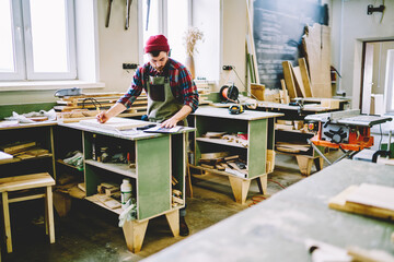 Skilled caucasian male artisan checking clients order while making furniture in manufacture...