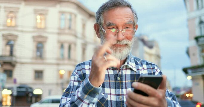 Caucasian old cheerful man with gray hair and beard, in glasses tapping and scrolling on mobile phone at street in town. Senior male pensioner texting message and typing on smartphone outdoors.