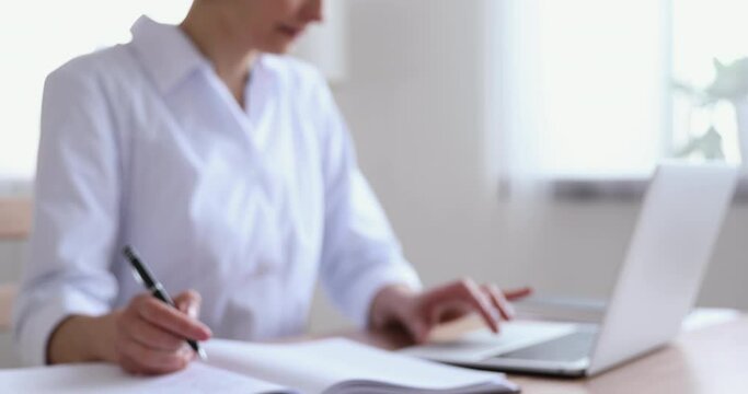 Focused young female general practitioner in white coat using medical software applications on computer, writing notes at workplace. Skilled professional nurse doctor consulting patient distantly.