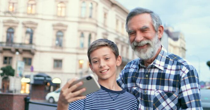 Caucasian teen happy boy taking selfie photo on smartphone camera together with senior grandfather with gray hair and beard at street in town. Grandpa and grandson posing to phone while making photos.