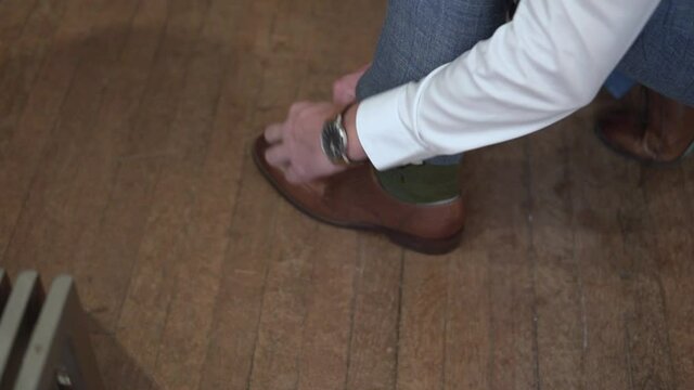 A man in blue trousers ties the laces on brown shoes standing on a wooden floor.