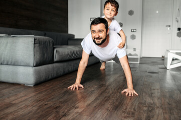 Sport at home with a child. Young dad does push-ups on the floor, his cute toddler son sits on top...
