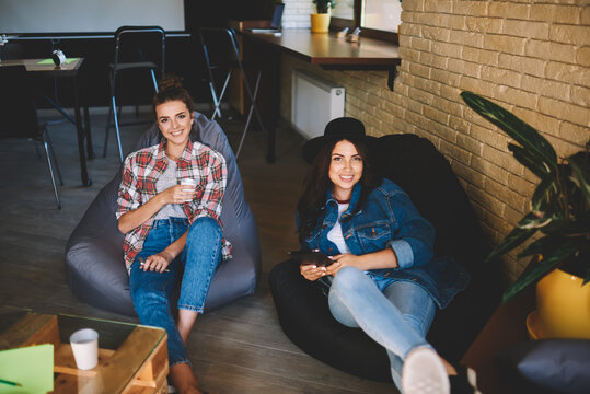 Portrait of two cheerful bloggers spending leisure time in stylish coworking space.Smiling hipster girls dressed in casual outfit looking at camera while relaxing during free time in office indoors