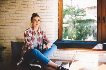 Portrait of positive hipster girl dressed in casual t-shirt sitting in coworking space and enjoying leisure time with laptop computer connecting to wireless 4G while looking at camera and smiling