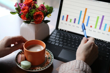 Home based remote work during new reality and quarantine, Social distancing concept.Working space view with cup of coffee with sweat treats between woman hands, working on the laptop computer and vase