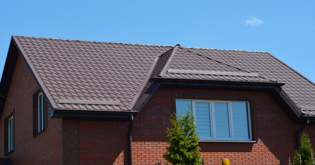 A close-up on a brown attic mansard metal tiled roof with snow guards, snow stoppers of a brick house against blue sky.