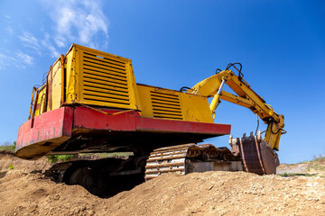 Obraz premium Yellow excavator during earthmoving at open pit on blue sky background. Construction machinery and earth-moving heavy equipment for excavation, loading, lifting and hauling of cargo on job sites