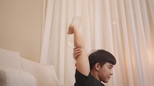 Asian young Man sit down back against couch doing arm workout with bottle of water as alternate dumbbell, home exercise, easy and convenience holding plastic water bottles triceps extension training