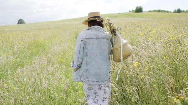 Mature woman sincerely smiling at camera, turned around ant started walking by the high green grass meadow with basket full of wildflowers. Human and nature concept slow motion footage.
