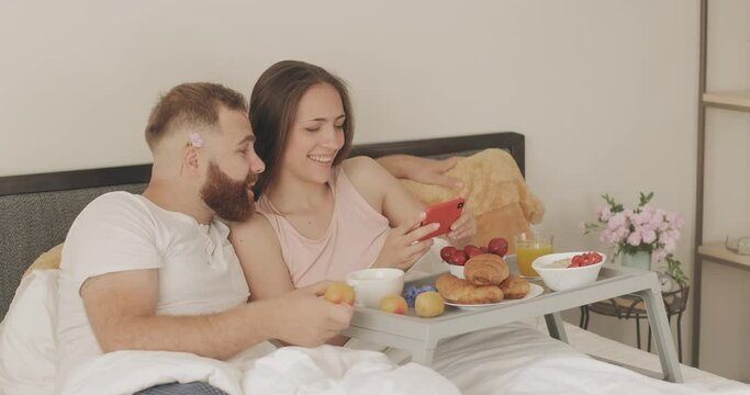 Cheerful young family talking and smiling while looking at phone screen. Man playing with fruit while woman holding smartphone during breakfast in bed. Concept of leisure.