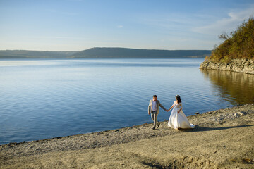 The bride and groom are walking near the lake on the shore.