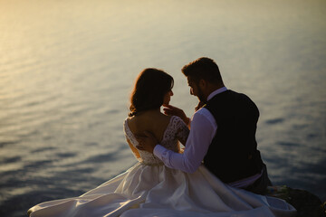 The bride and groom are sitting on the edge of a cliff against the backdrop of the lake.