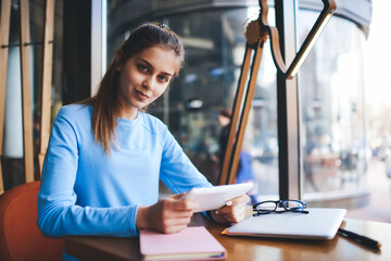 Portrait of beautiful female blogger in good mood looking at camera while messaging with followers in personal website on touchpad connected to 4G internet sitting at wooden table