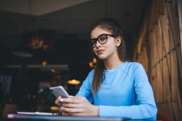 Portrait of attractive female blogger in stylish eyeglasses for good vision texting new article for personal web page on smartphone.Good looking woman sending sms to friends using application