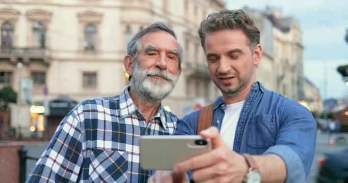 Caucasian happy handsome man taking selfie photo on smartphone camera together with senior gray-haired father at street in city. Cheerful old dad and son posing to phone while making photos outside.