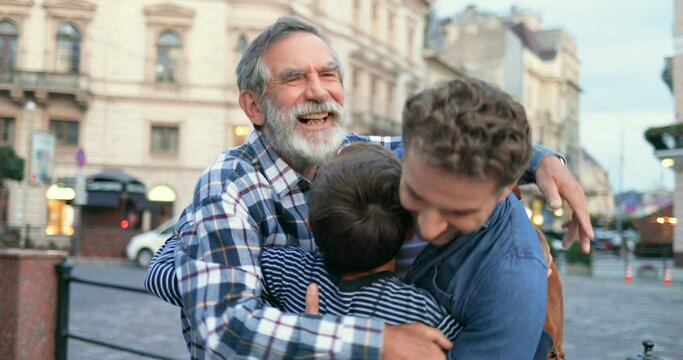 Two Caucasian men standing outdoor and talking. Caucasian teen boy running to handsome father and senior grandfather and they hugging happily at steet in town. Son coming and embracing grandpa and dad