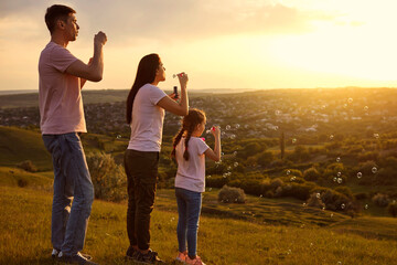 Affectionate family having fun blowing soap bubbles on mountain top at sunset. Parents and child playing together